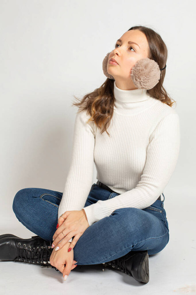 Looking up young woman in warm white jumper with fur earmuffs isolated on white sitting on the floor with crossed legs in jeans - Zdjęcie, obraz
