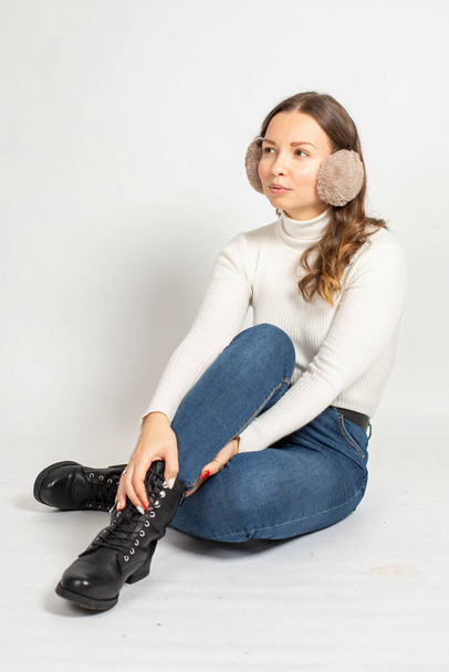 Young woman in warm white jumper with fur earmuffs isolated on white with crossed legs on the floor - Foto, imagen