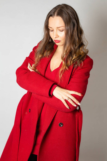 Attractive young european woman in red coat isolated on light grey with folded arms - Фото, зображення