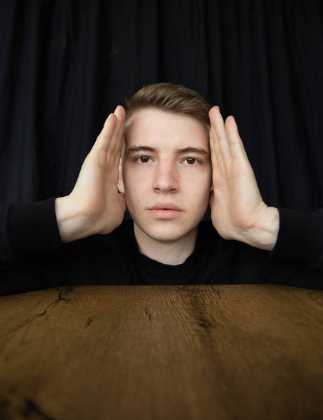Portrait of young blond man isolated on black holding hands near his face above the table - Photo, image