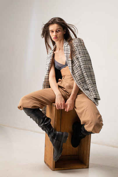 Young slim woman in studio sitting on wooden crate with plaid blazer on beige - Foto, Imagem