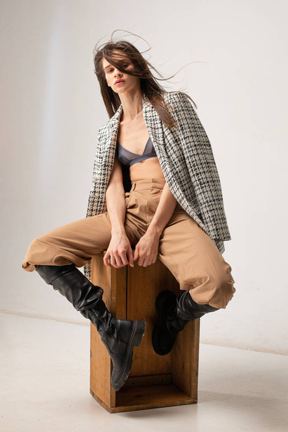 Young slim woman in studio with plaid blazer sitting on crate on beige - Fotografie, Obrázek