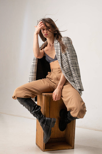 Young slim woman in studio with plaid blazer sitting on crate on beige - Foto, imagen