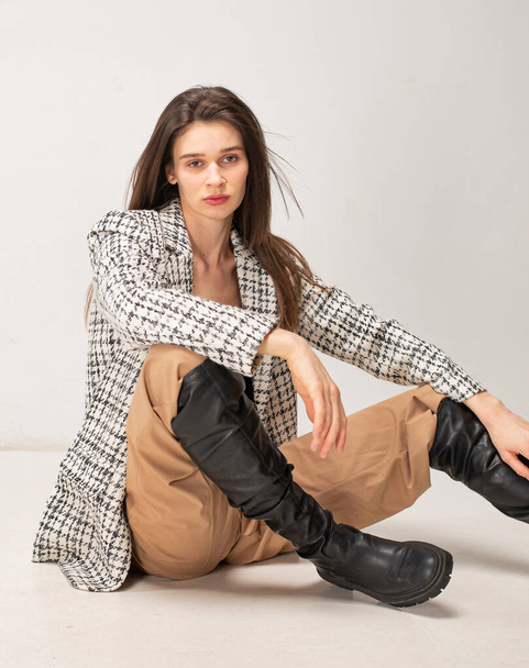 Young slim woman with dark hair isolated on light beige sitting on the floor in plaid blazer - Fotó, kép