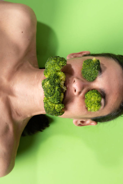 Girl lying down with broccoli in eyes and mouth on green background. - Фото, зображення