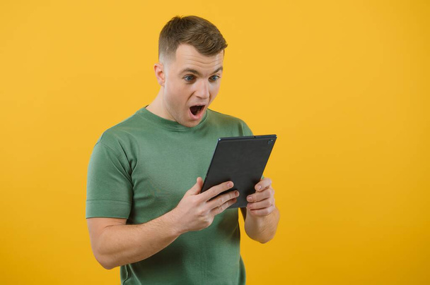 Amazed happy man using digital tablet looking shocked about social media news, astonished man shopper consumer surprised excited by online win isolated over yellow background. - Фото, изображение