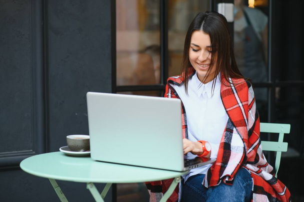 Young freelancer woman enjoying coffee while working outdoors on a laptop computer connected to public Wi-Fi. Stylish student girl studying online in city. - Фото, зображення