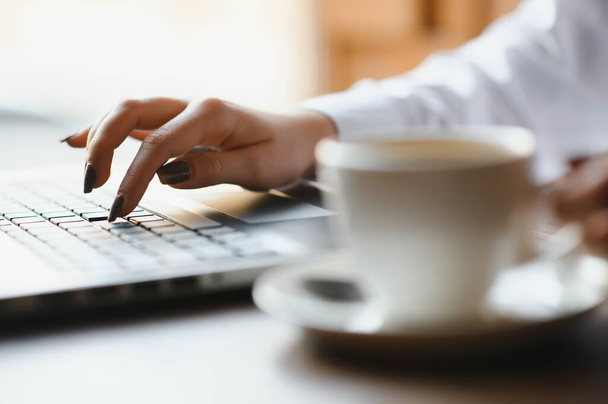Nahaufnahme Hände einer Geschäftsfrau in pastellrosa Hemd, die am Laptop arbeitet und Notizen in ihrem Notizbuch auf einem Holztisch mit einer Tasse Kaffee macht - Foto, Bild