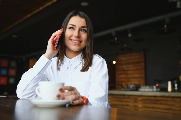 Young charming woman calling with cell telephone while sitting alone in coffee shop during free time, attractive female with cute smile having talking conversation with mobile phone while rest in cafe - Foto, Bild