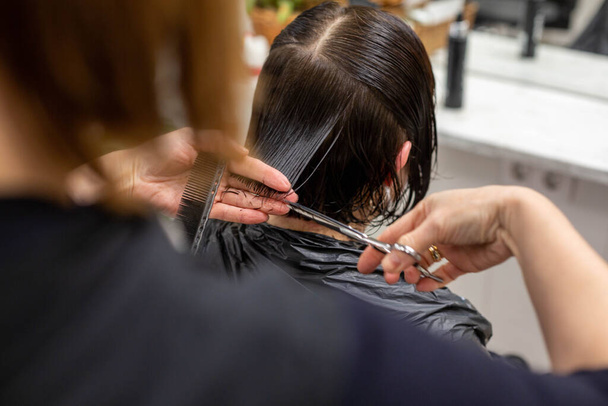 Una peluquera corta el pelo de una mujer morena en un salón de belleza. Tema de belleza y cuidado del cabello. - Foto, Imagen