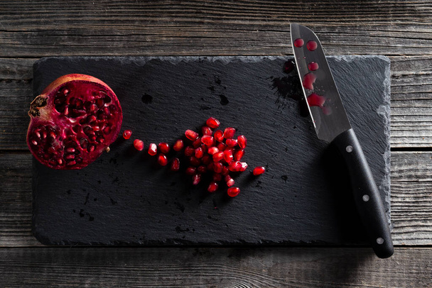 Eine halbe rote Granatapfelfrucht und ein Stapel Kerne auf einem schwarzen Schieferschneidebrett mit einem Küchenmesser auf einem hölzernen Tischhintergrund. Das Konzept der gesunden Ernährung, Ernährung. - Foto, Bild