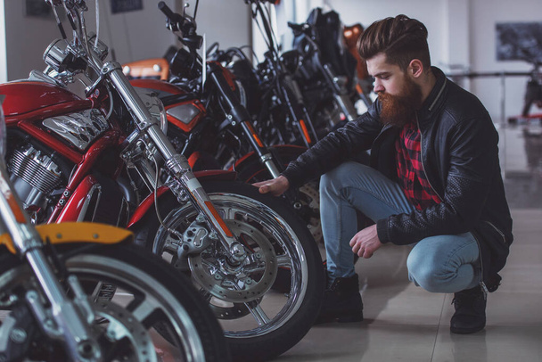 Красивый молодой бородатый мужчина в черной кожаной куртке осматривает мотоциклы в мотосалоне
 - Фото, изображение