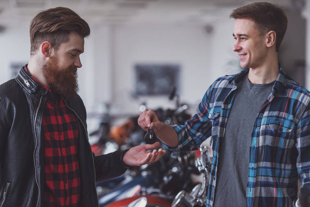 Handsome young bearded man in black leather jacket is buying a motorbike, attractive blond man is giving keys - Photo, Image