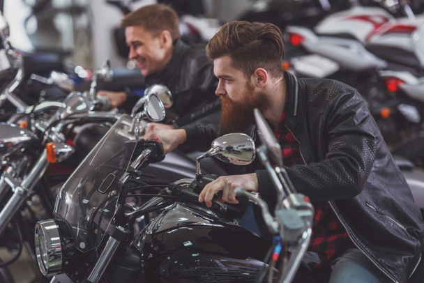 Handsome young bearded and blond men in black leather jackets are imitating ride and smiling while sitting on motorbikes - Photo, Image
