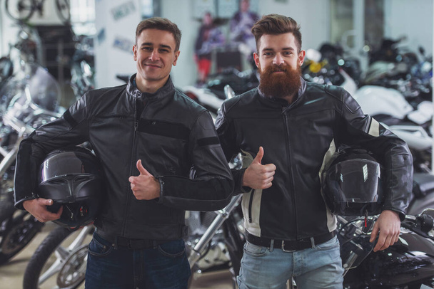 Handsome young bearded and blond men in black leather jackets are holding helmets, showing Ok sign and smiling while standing in motorbike salon - Photo, Image
