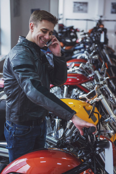 Attractive young blond man in black leather jacket is talking on the mobile phone and smiling while standing in a motorbike salon - Photo, Image