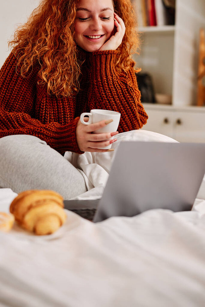 A happy ginger girl sits on the bed in the morning, drinking her morning coffee and using her laptop. - Photo, image