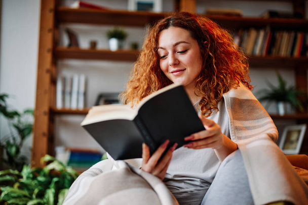 A beautiful ginger girl sitting in the living room wrapped in a blanket and reading an interesting book in the morning. - Fotografie, Obrázek