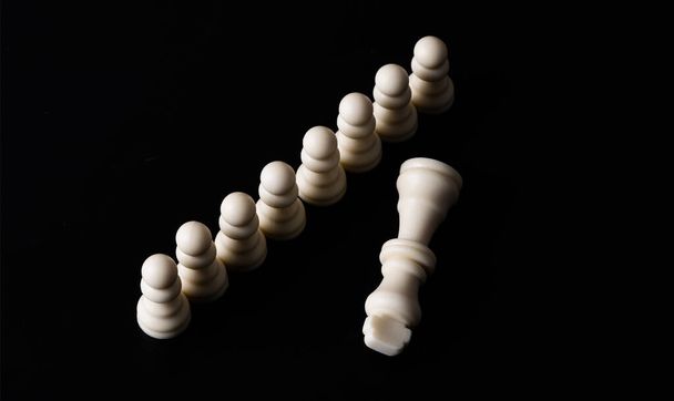 The defeated figure of the chess king in front of the chess pawns on a black background - Photo, Image