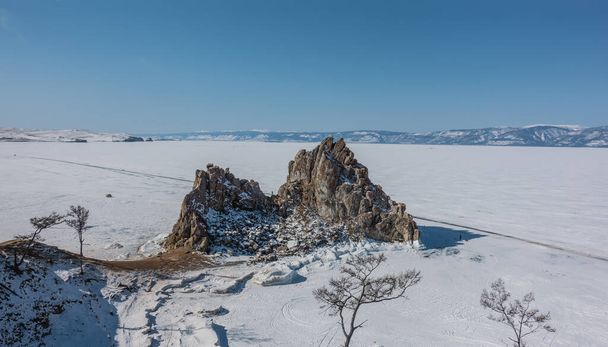 Malownicza dwugłowa granitowa skała na tle zamarzniętego jeziora. Śnieg na stromych stokach i na lodzie. Gołe drzewa na pierwszym planie. Błękitne niebo. Bajkał. Góra Shamanka - Zdjęcie, obraz