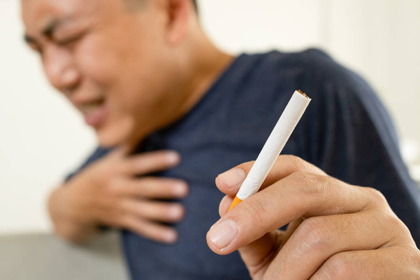 Ασιάτης καπνιστής κρατά ένα τσιγάρο, αγγίξτε τον πόνο στο στήθος με το χέρι, δυσκολία στην αναπνοή, ασφυξία από το κάπνισμα, άνθρωπος που κρατά το στήθος του με οξεία δύσπνοια, καρδιακή προσβολή, ασθένειες της καρδιάς ή των πνευμόνων έννοια - Φωτογραφία, εικόνα