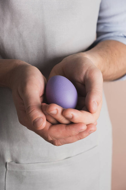 Las manos del hombre sostienen huevo de Pascua pintado en color púrpura. Preparación para la Pascua, cocinar en la cocina casera. Primer plano, vertical - Foto, Imagen