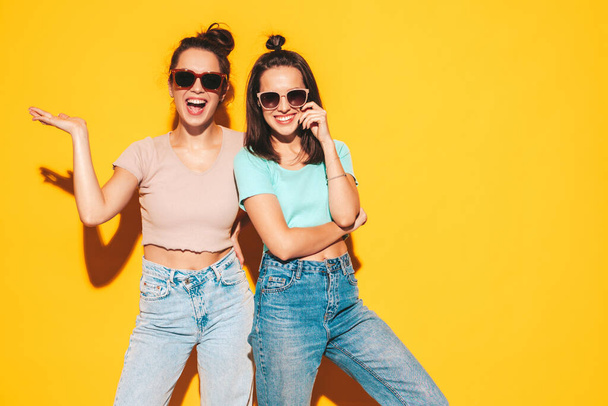 Two young beautiful smiling blond hipster female in trendy summer clothes. Sexy carefree women posing near yellow wall in studio. Positive models having fun. Cheerful and happy. In sunglasses - Foto, Imagen