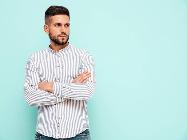 Portrait of handsome confident model. Sexy stylish man dressed in shirt and jeans. Fashion hipster male posing near blue wall in studio. Crossed arms - 写真・画像