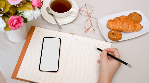 Primer plano, imagen recortada, Una mujer escribe algo en su diario con un lápiz y un teléfono inteligente en la mesa de café. - Foto, imagen