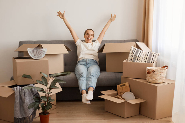 Portrait of excited positive woman wearing white casual T-shirt and jeans, sitting on sofa, surrounded with boxes during moving, looking at camera with raised arms, celebrating buying new flat. - Foto, Imagem
