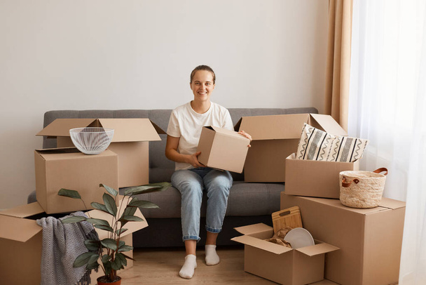 Indoor shot of optimistic good looking woman wearing white casual T-shirt and jeans, sitting on sofa, surrounded with boxes during relocating,holding package with personal pile, looks happy. - Fotografie, Obrázek
