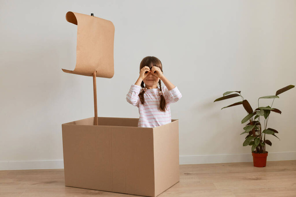 Indoor shot of cute funny little girl sitting in cardboard box with paper flag, playing ship captain, pretending she is in the sea, making binoculars with her hand and looking at camera. - Photo, image