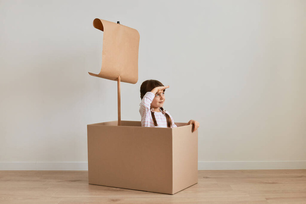 Portrait of attentive little girl sitting in cardboard box with paper flag, playing ship captain, keeping hand on her forehead, looking far, pretending she is in the sea. - Foto, Imagem