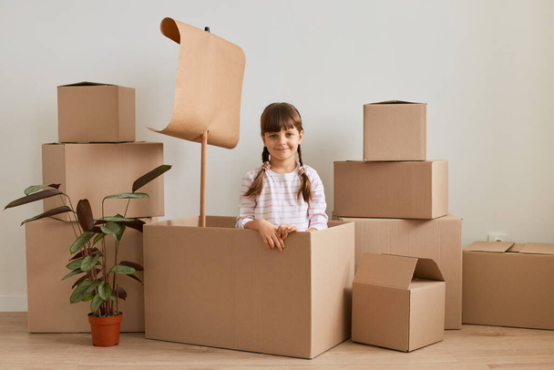 Portrait of smiling little girl making ship from carton box with flag during relocation and playing sailor at new house, looking at camera with happy facial expression, glad to move into new flat. - Φωτογραφία, εικόνα