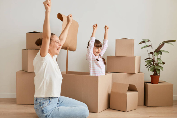 Indoor shot of young adult woman with little girl making ship with flag during relocation and playing sailor at new apartment, celebrating moving, posing with raised arms and happy expression. - Photo, Image