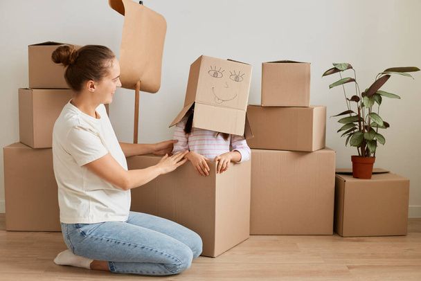 Portrait of woman in white t shirt and jeans sitting near packages with personal belongings and playing with her daughter sitting with cardboard box with drawn positive face on her head. - Foto, imagen