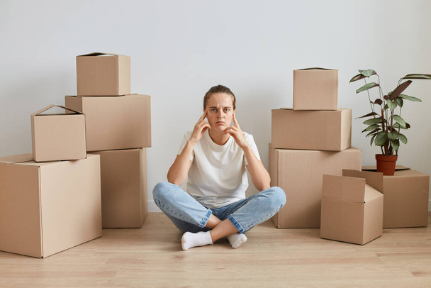 Indoor shot of tired pensive woman wearing white casual style t shirt, sitting on floor with crossed legs surrounded with cardboard packages, keeping hands on head, being thoughtful. - Фото, изображение