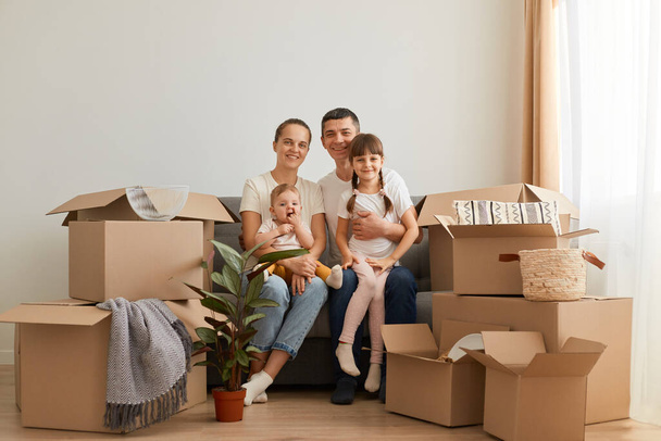 Portrait of happy family with children sitting on cough surrounded with cardboard boxes with personal piles, unpacking packages after moving in to new apartment. - Zdjęcie, obraz