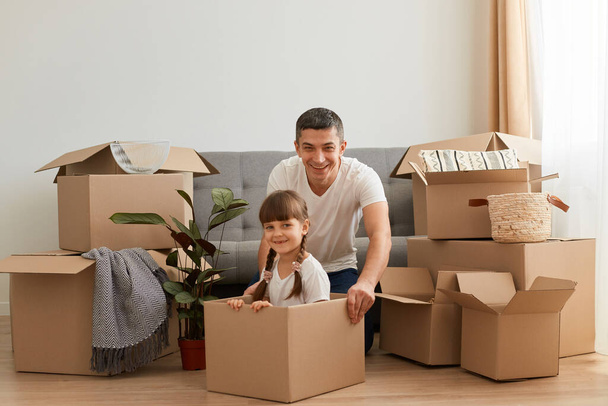 Father playing with his daughter in their new home, riding in a cardboard box laughing, all seem ecstatic and overfilled with positive emotions, relocating to a new apartment. - Foto, afbeelding