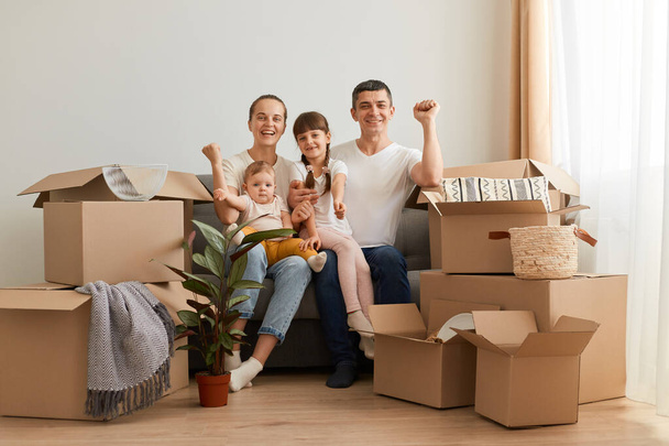 Happy family sitting on cough in a new apartment, celebrating relocation, clenched fists, moving to a new big light house, being surrounded with cardboard packages. - Фото, зображення