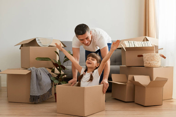 Indoor shot of handsome brunette man riding in a cardboard box his excited daughter, laughing, little girl raised arms, celebrating moving to a new flat. - Valokuva, kuva