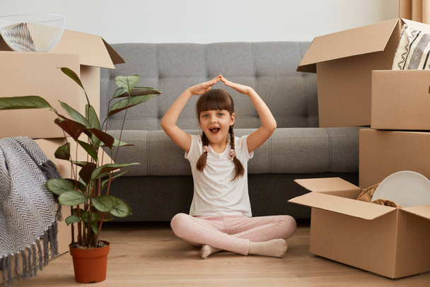 Photo of little girl sitting on floor and making roof above head, feeling safety, satisfied relocation to a new house, looking at camera with open mouth and excited facial expression. - 写真・画像