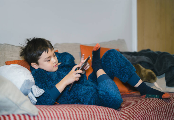 Cinematic portrait happy kid watching cartoon on tablet, Candid shot cute boy playing game online with friends on internet, Cozy Child wearing fluffy pajamas sitting on sofa relaxing at home  - Foto, Imagen