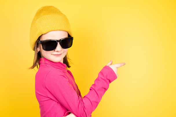 Little girl on winter clothes and sunglasses pointing to copy space while looking to camera. - Φωτογραφία, εικόνα