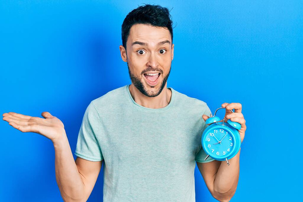 Young hispanic man holding alarm clock celebrating achievement with happy smile and winner expression with raised hand  - Foto, imagen