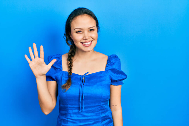 Fiatal spanyol lány hétköznapi ruhákat visel, mutatva és mutatva az ötös számú ujjaival, miközben magabiztosan és boldogan mosolyog..  - Fotó, kép