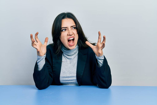 Piękna latynoska bizneswoman siedząca na stole szalona i szalona krzycząca i wrzeszcząca z agresywnym wyrazem twarzy i podniesionymi ramionami. koncepcja frustracji.  - Zdjęcie, obraz