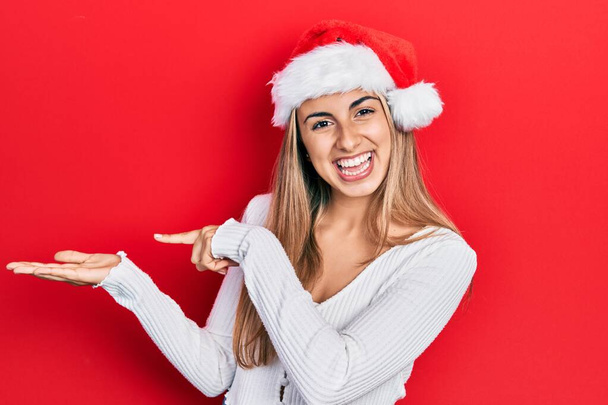 手で提示し、指で指摘しながら、クリスマスの帽子を身に着けている美しいパニック女性は驚きとカメラに笑みを浮かべて.  - 写真・画像
