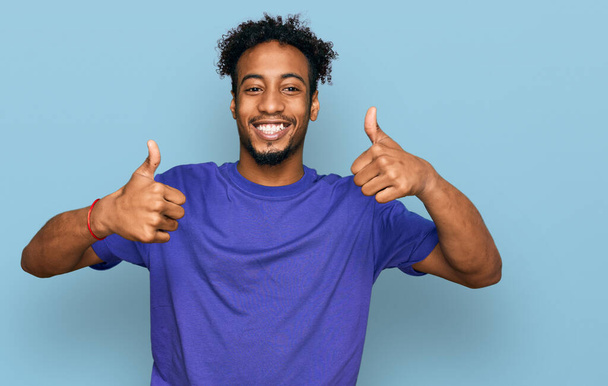 Ein junger afrikanisch-amerikanischer Mann mit Bart trägt ein legeres lila T-Shirt und macht eine positive Geste mit der Hand, Daumen hoch lächelnd und glücklich. Fröhlicher Ausdruck und Siegergeste.  - Foto, Bild