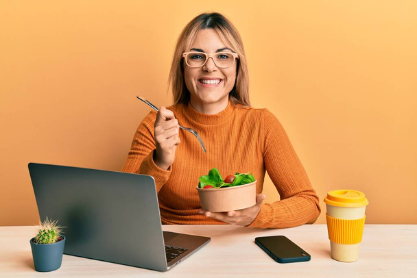 Mujer caucásica joven que trabaja en la oficina comiendo ensalada saludable sonriendo con una sonrisa alegre y fresca en la cara. mostrando dientes.  - Foto, Imagen